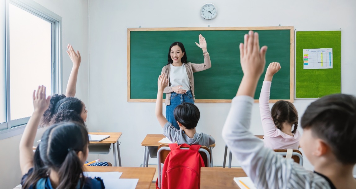 A female teacher standing in front of a blackboard while several students raise their hands to answer a question in a classroom.